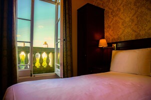 A hotel room with a bed, a nightstand with a lamp, a window with curtains, and a view of a building outside.