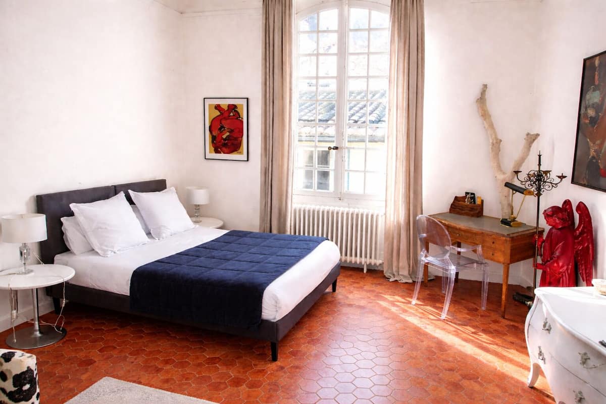 A hotel room with a bed, a desk, a chair, a radiator, and a window with curtains.