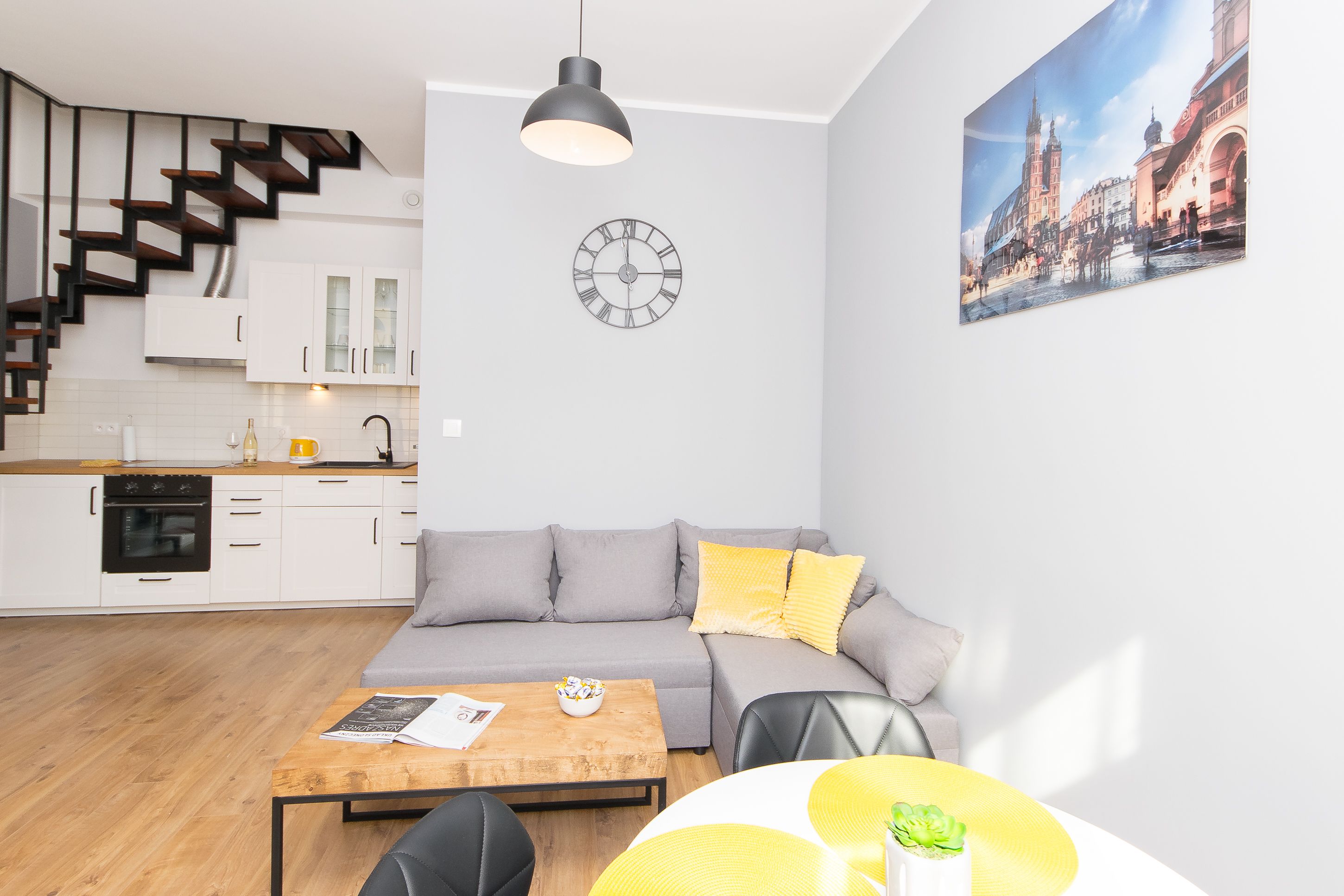 A modern apartment with a kitchen, a gray sofa with yellow pillows, a wooden coffee table, and a large clock on the wall.