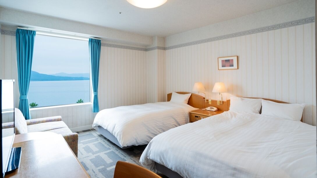 A hotel room with two beds, a desk, a chair, a window with curtains, and a lamp on the nightstand.