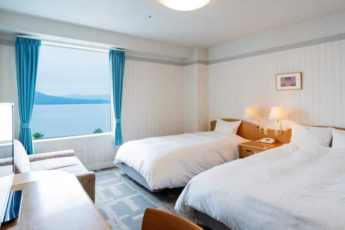 A hotel room with two beds, a desk, a chair, a window with curtains, and a lamp on the nightstand.