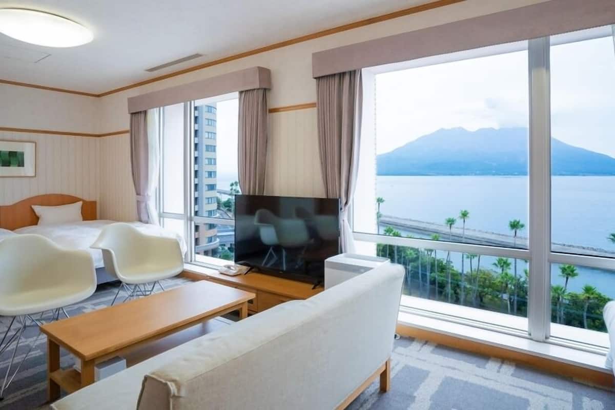 A hotel room with a bed, a TV, a sofa, a coffee table, and large windows with a view of a body of water and mountains.