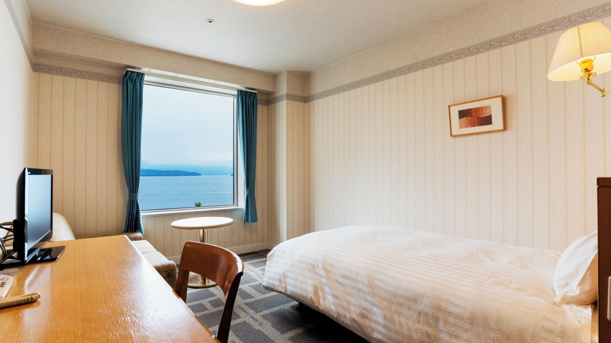 A hotel room with a bed, a desk with a computer, a chair, a small table, a window with curtains, and a wall-mounted lamp.