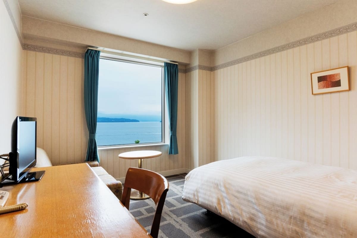A hotel room with a bed, a desk with a computer, a chair, a small table, a window with curtains, and a wall-mounted lamp.