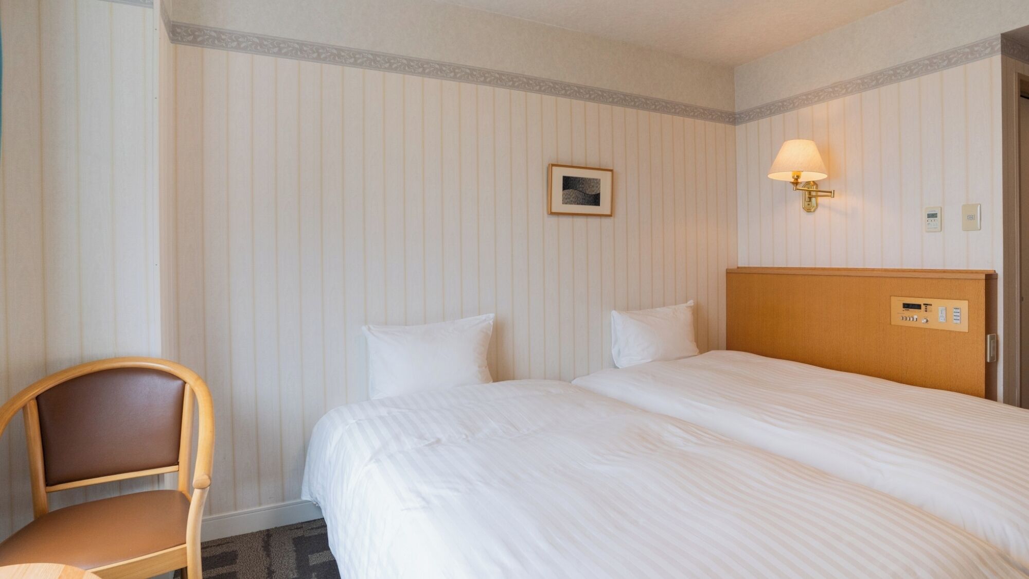 A hotel room with a bed, a chair, a wall-mounted lamp, and a small picture frame.