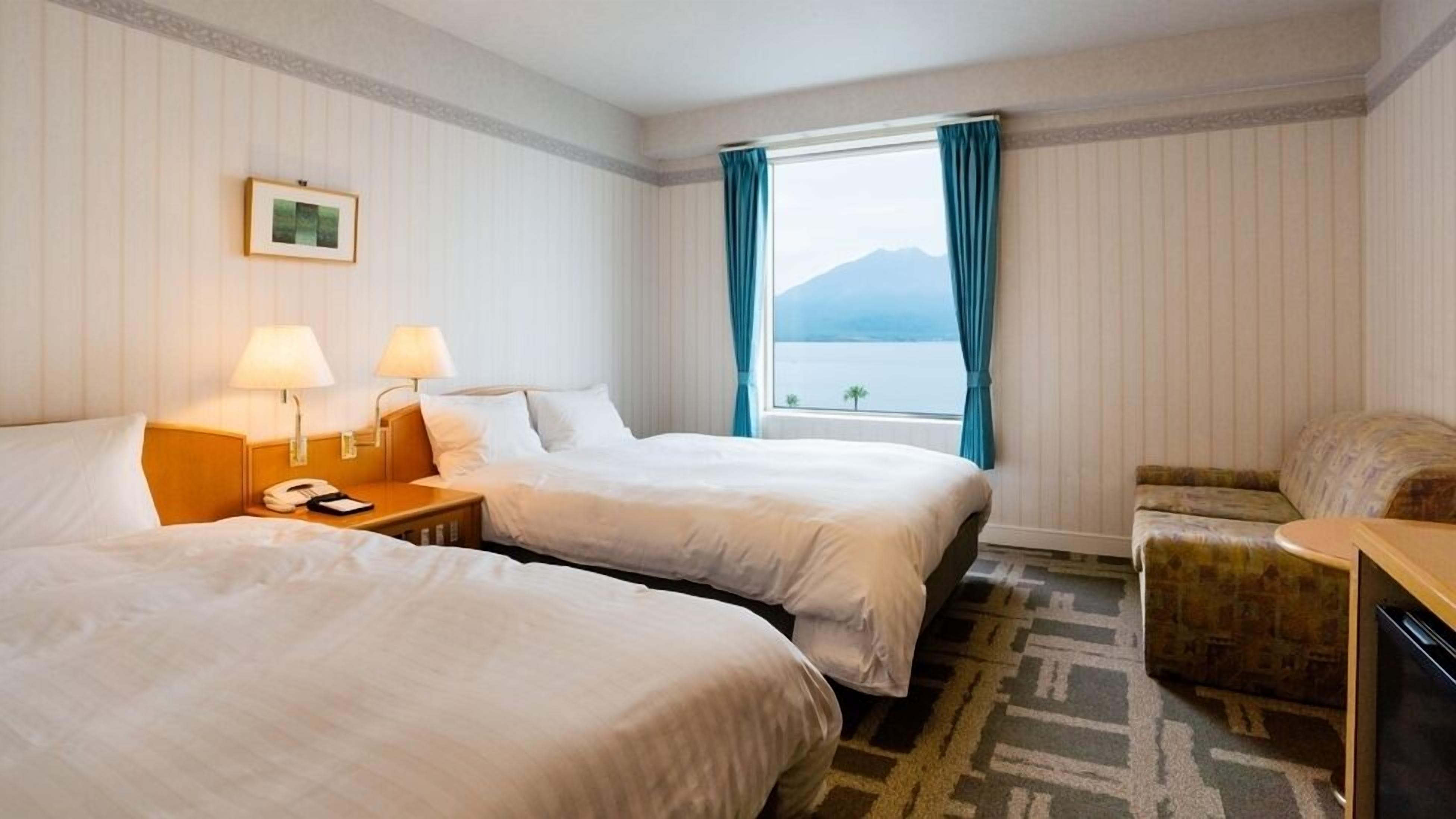A hotel room with two beds, a desk, a chair, a window with a view of the sea, and a framed picture on the wall.