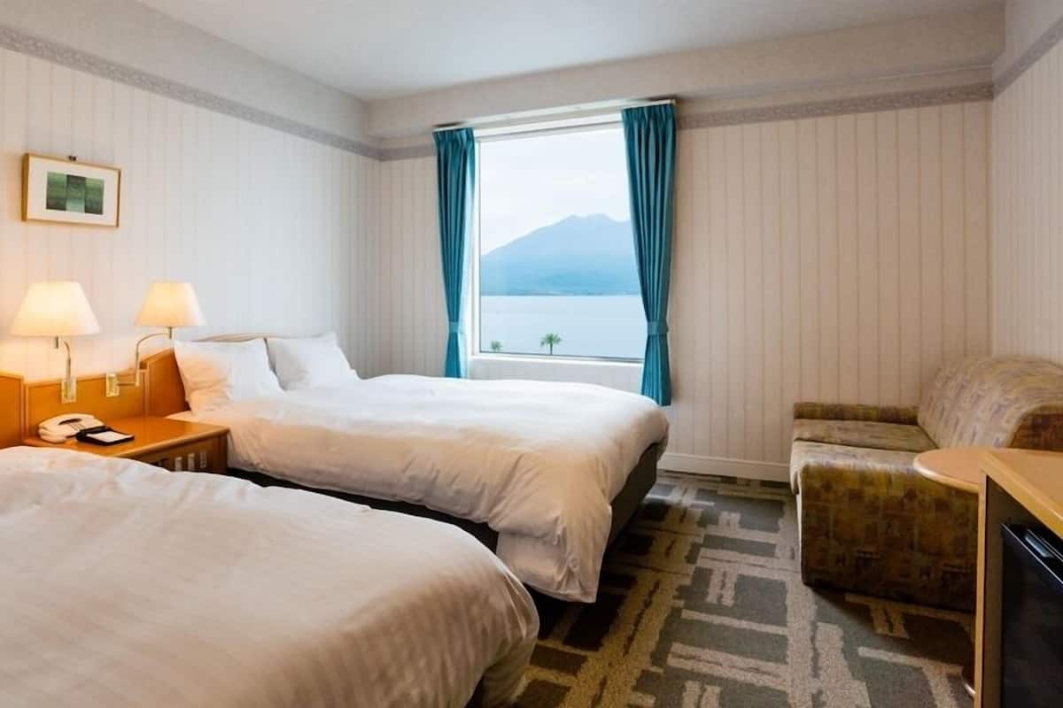 A hotel room with two beds, a desk, a chair, a window with a view of the sea, and a framed picture on the wall.