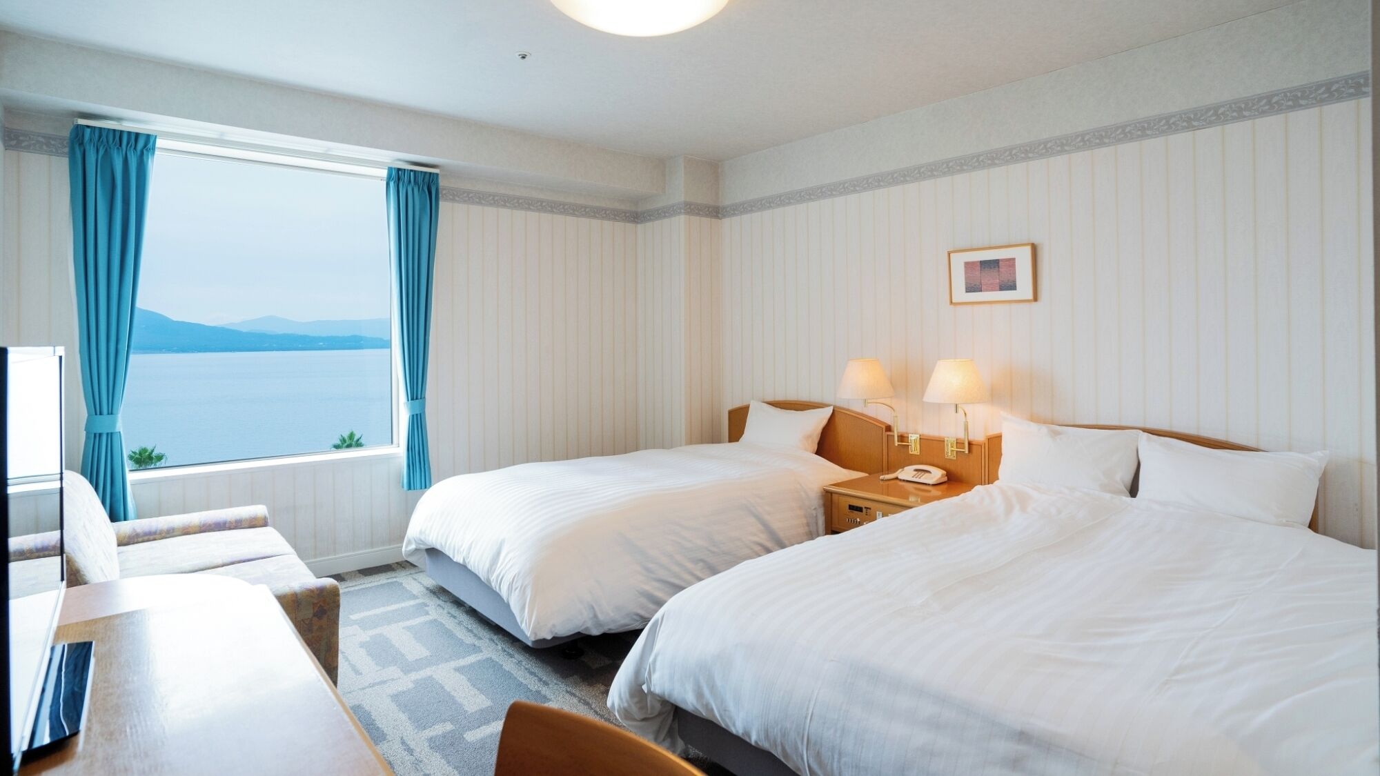 A hotel room with two beds, a desk, and a view of the sea.