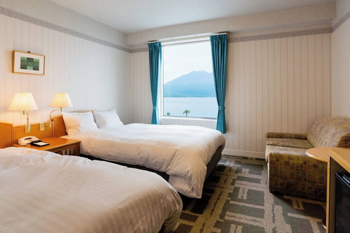 A hotel room with two beds, a desk with a lamp, a chair, and a view of the sea.