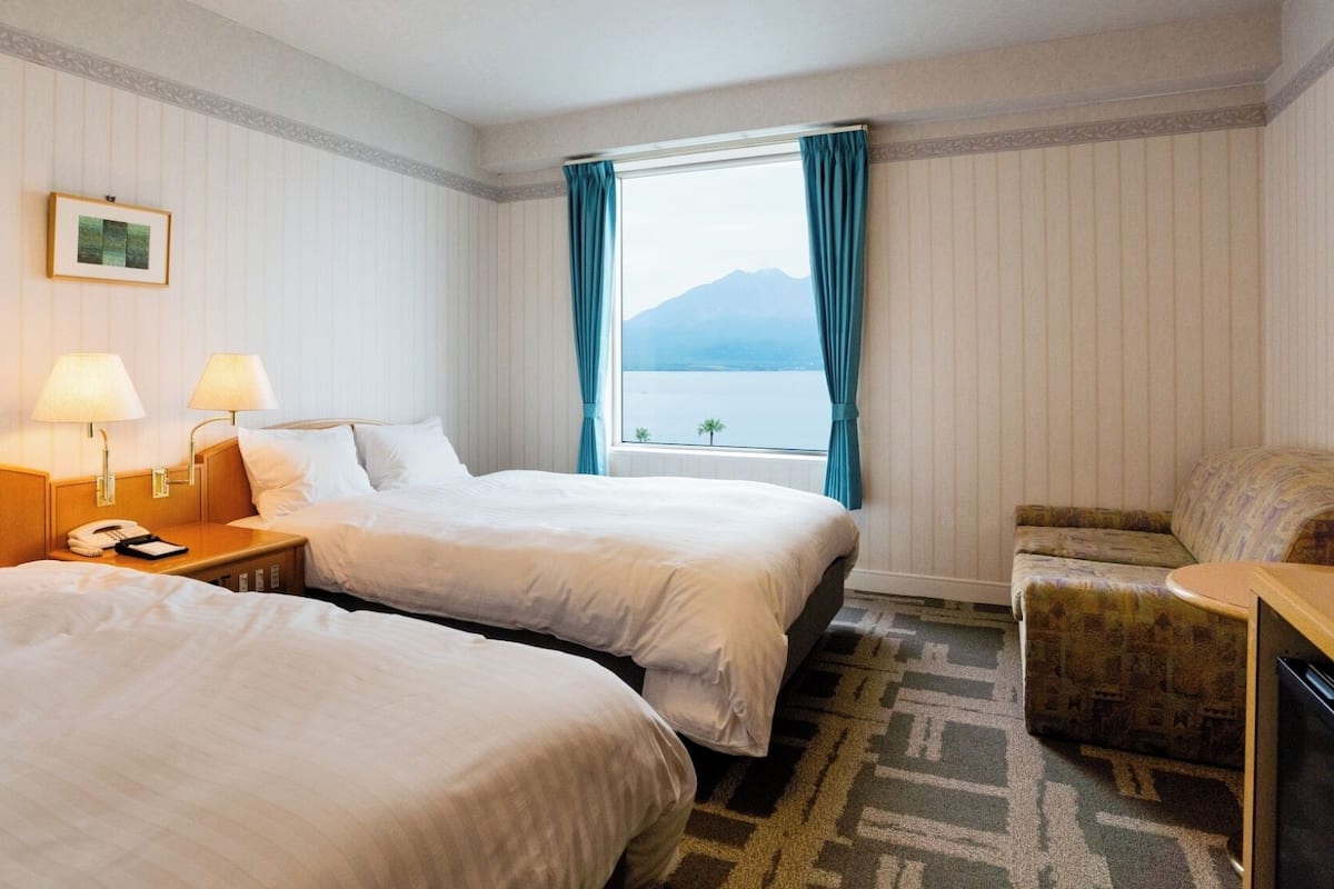 A hotel room with two beds, a desk with a lamp, a chair, and a view of the sea.