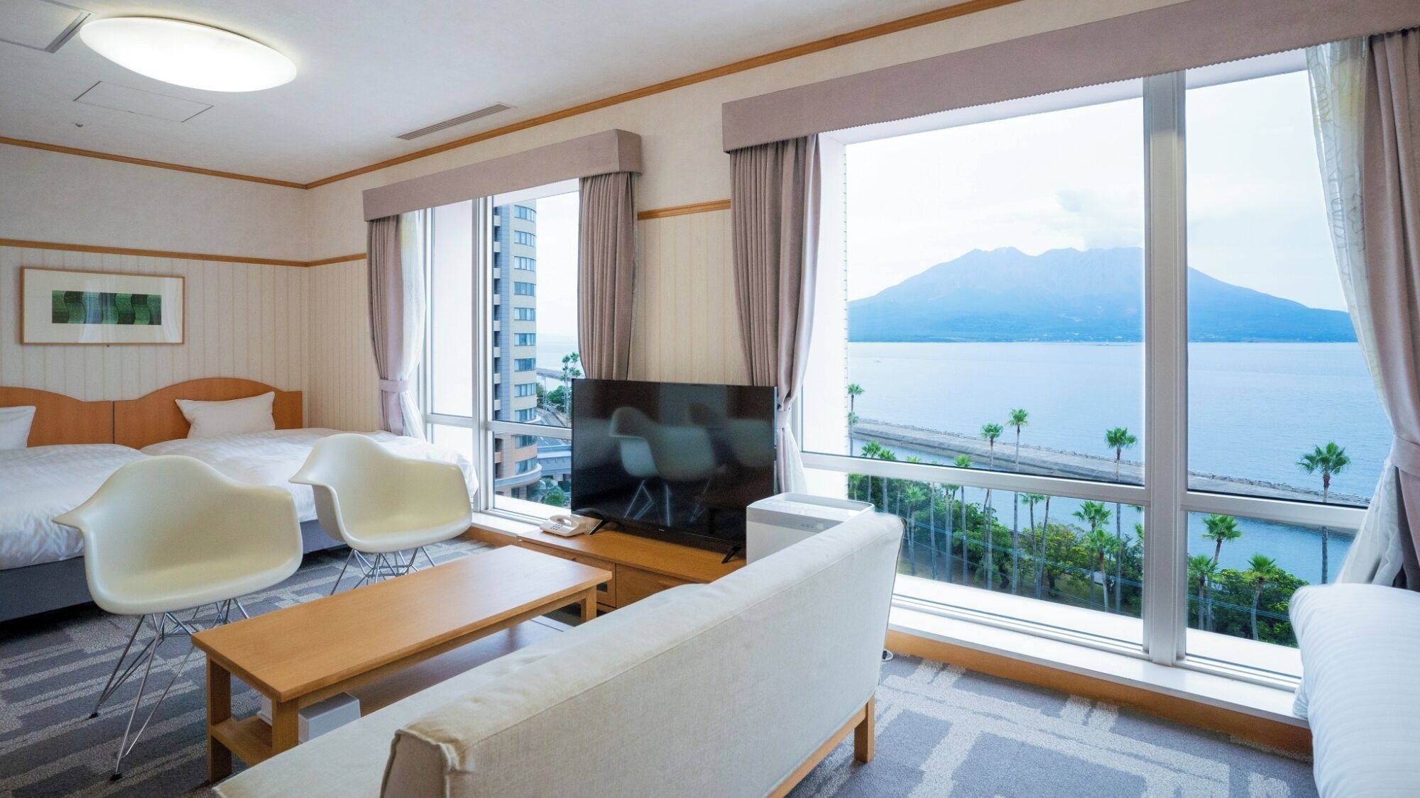 A hotel room with a bed, a TV, a sofa, a coffee table, and a large window with a view of a body of water and mountains.