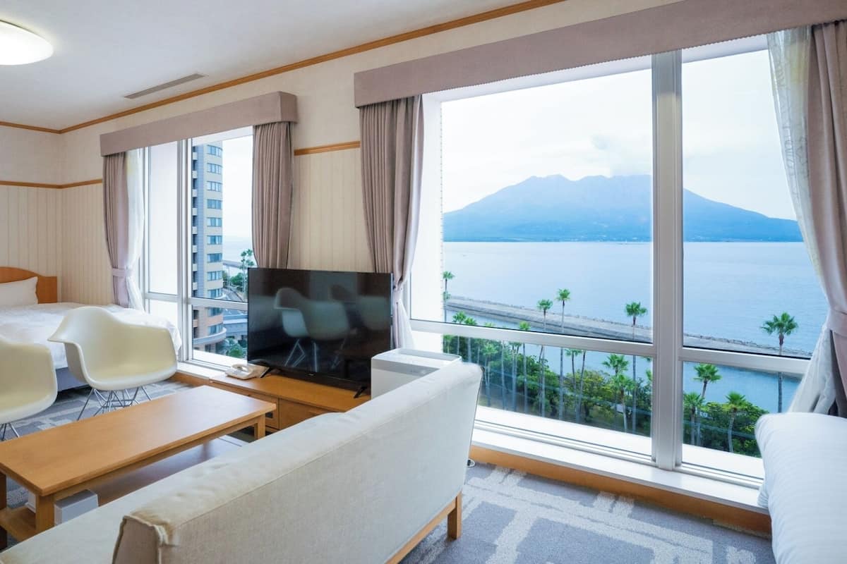A hotel room with a bed, a TV, a sofa, a coffee table, and a large window with a view of a body of water and mountains.