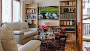 Una sala de estar con un sofá color crema, una mesa de centro de vidrio con un jarrón de flores, una estantería y un televisor mostrando una escena de película.