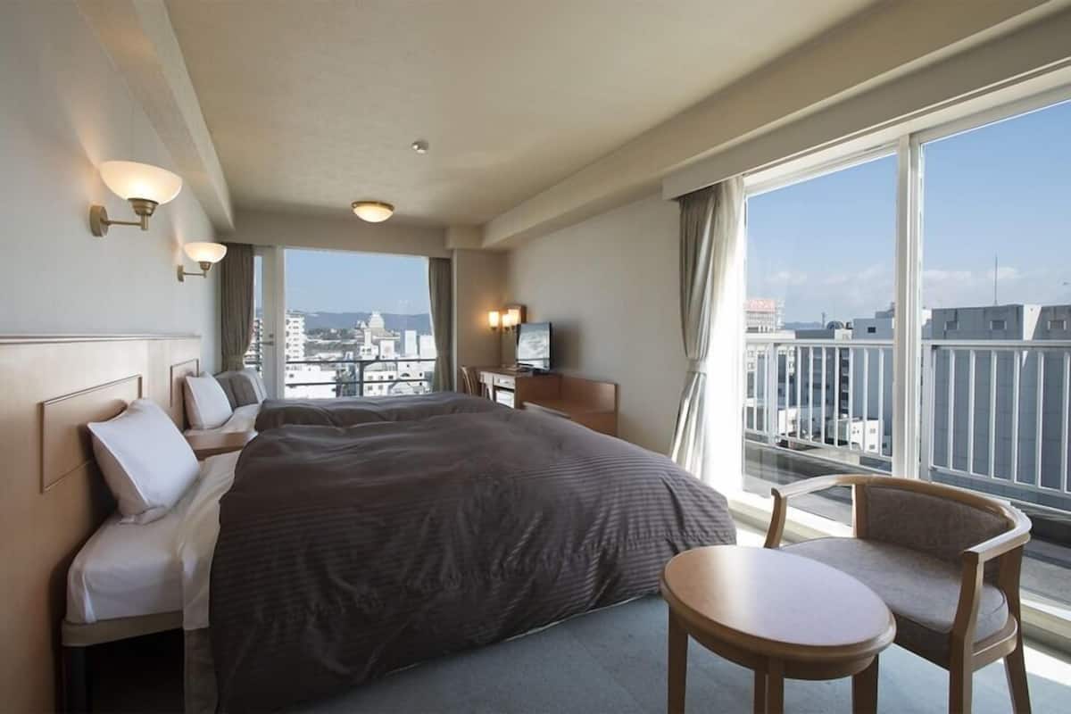 Een hotelkamer met een groot bed, een bureau, een stoel en uitzicht op de stad door grote ramen.