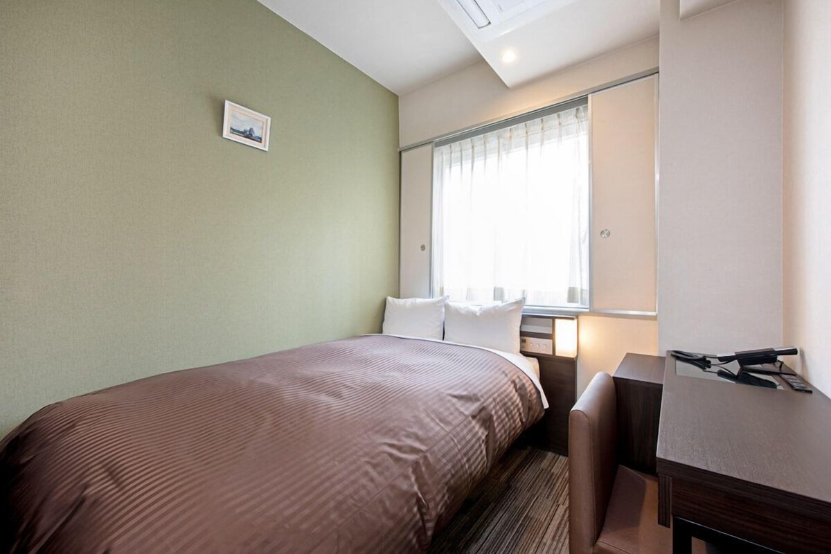 Ein Hotelzimmer mit Bett, Schreibtisch, Stuhl, einem Fenster mit Vorhängen und einem Bild an der Wand.