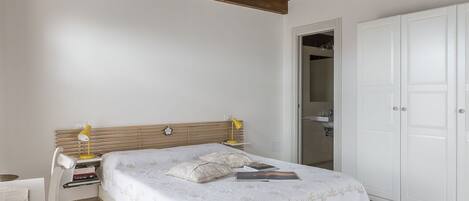 Un dormitorio con estructura de cama de madera, ropa de cama blanca, una mesita de noche con lámpara y un armario.