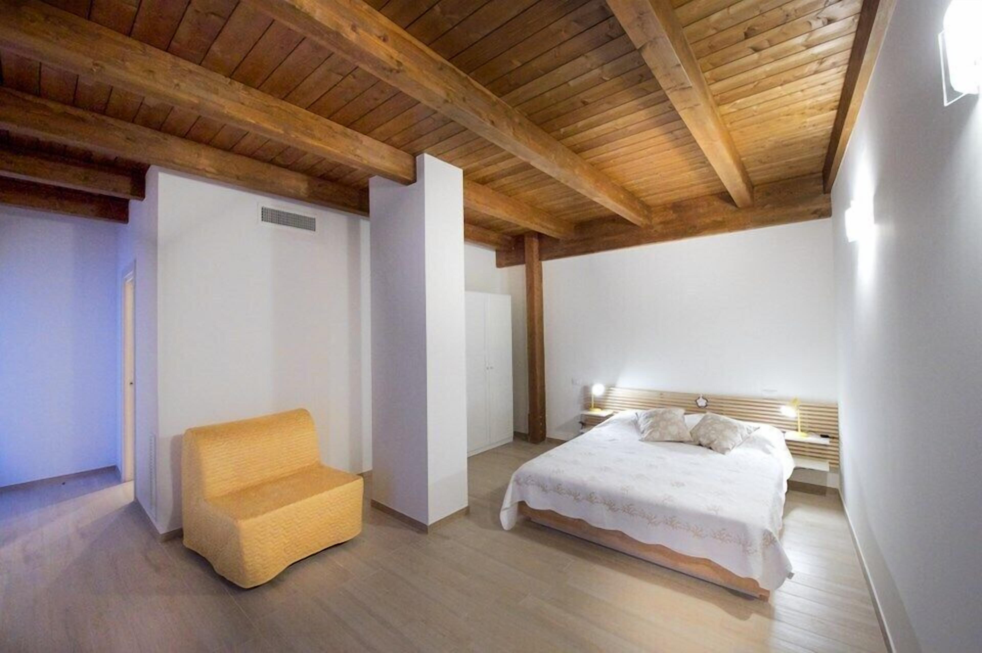 A bedroom with a wooden ceiling, a bed with a white bedspread, a nightstand, and a yellow chair.