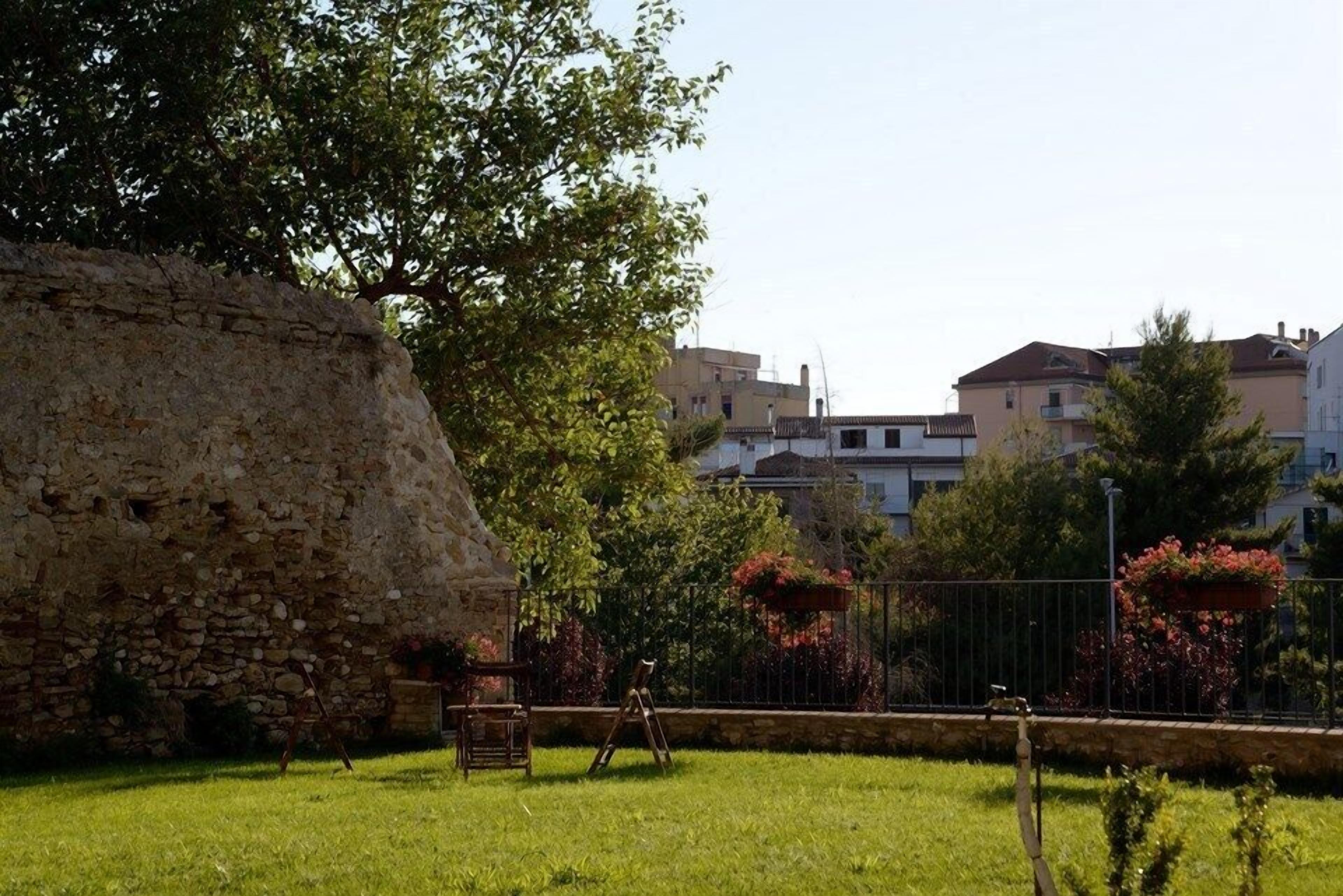 Un muro de piedra, un espacio con césped y un edificio con balcones al fondo.
