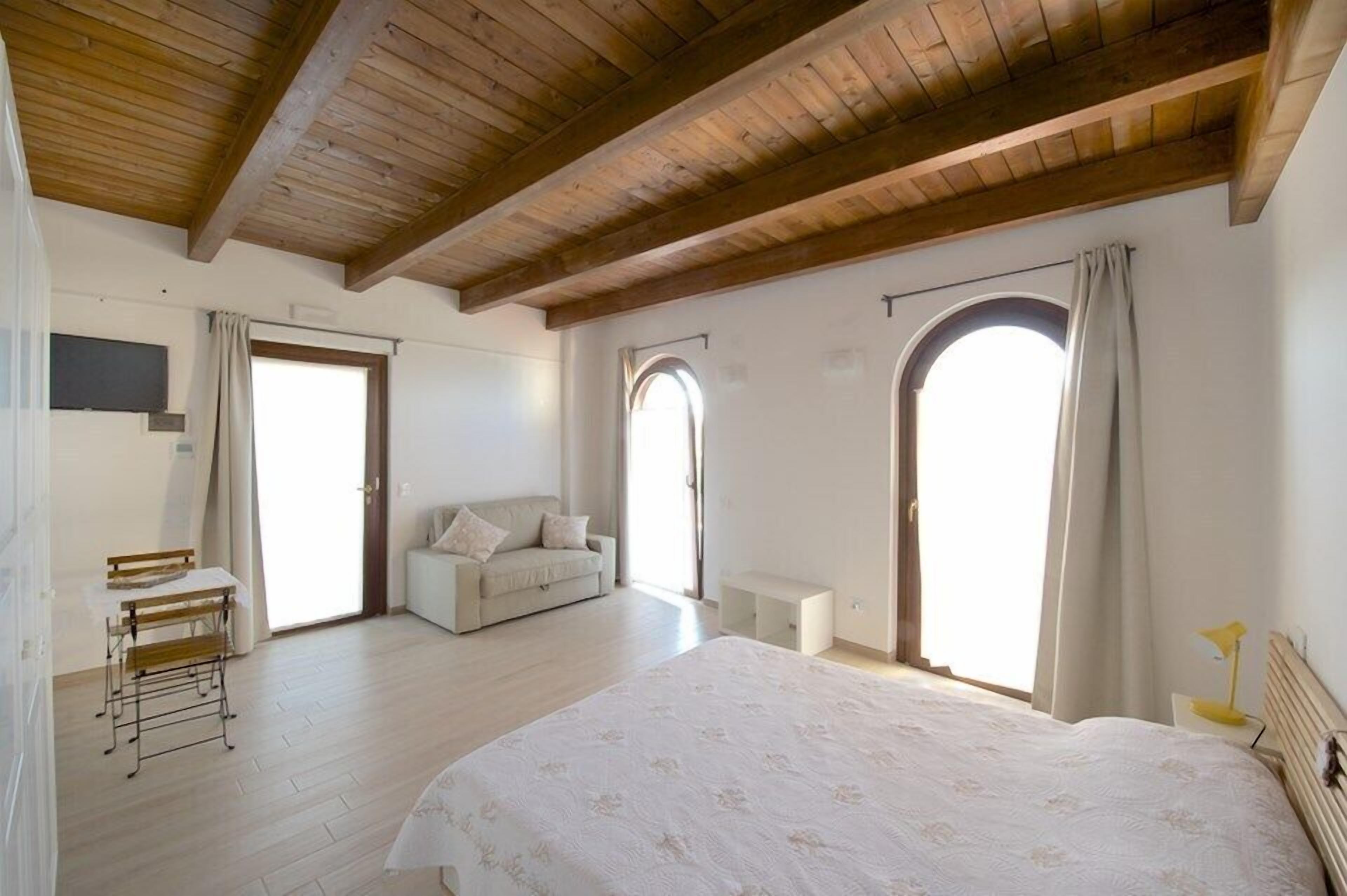 Um quarto espaçoso com uma cama grande, um sofá e uma escrivaninha. Há duas janelas em arco e teto de madeira.