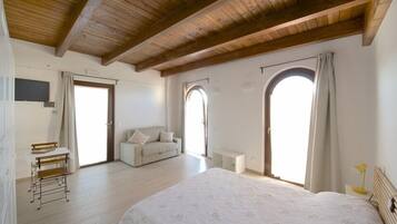 Um quarto espaçoso com uma cama grande, um sofá e uma escrivaninha. Há duas janelas em arco e teto de madeira.