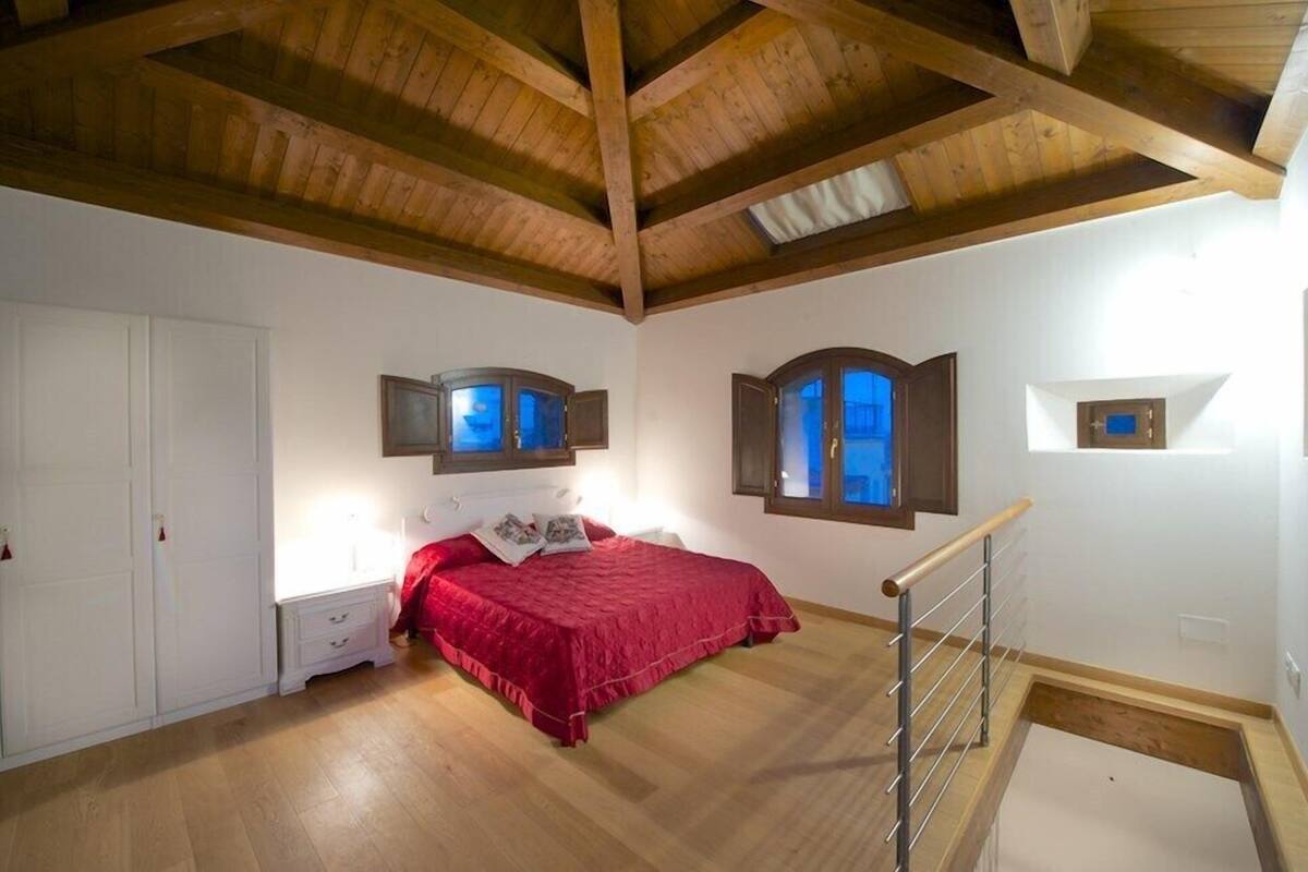 Une chambre avec un plafond en bois, un lit recouvert d’une couverture rouge, une table de chevet et une fenêtre donnant sur un beau paysage.