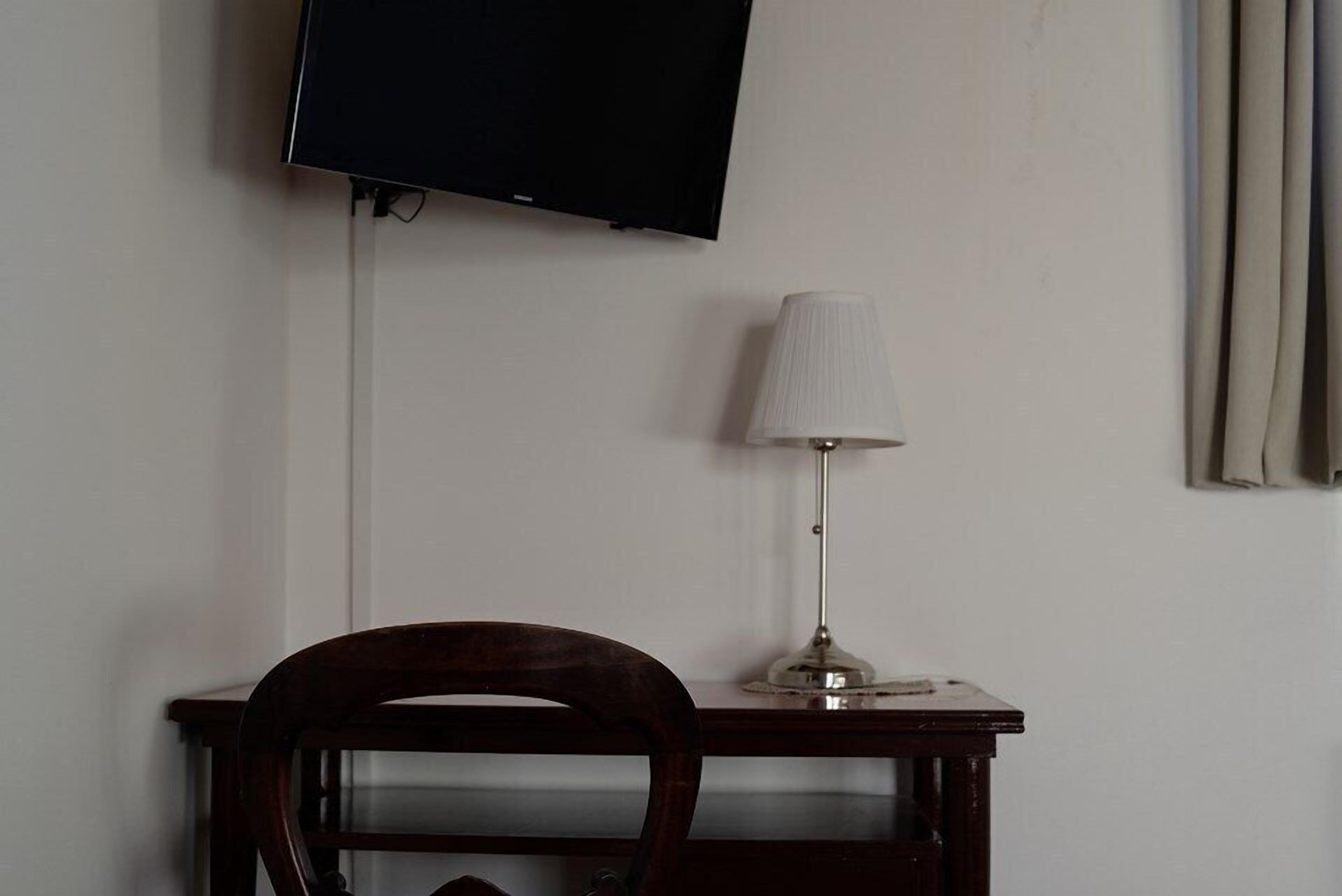 Ein Zimmer mit einem an der Wand montierten Flachbildfernseher, einem Schreibtisch mit Lampe und einem Sessel.