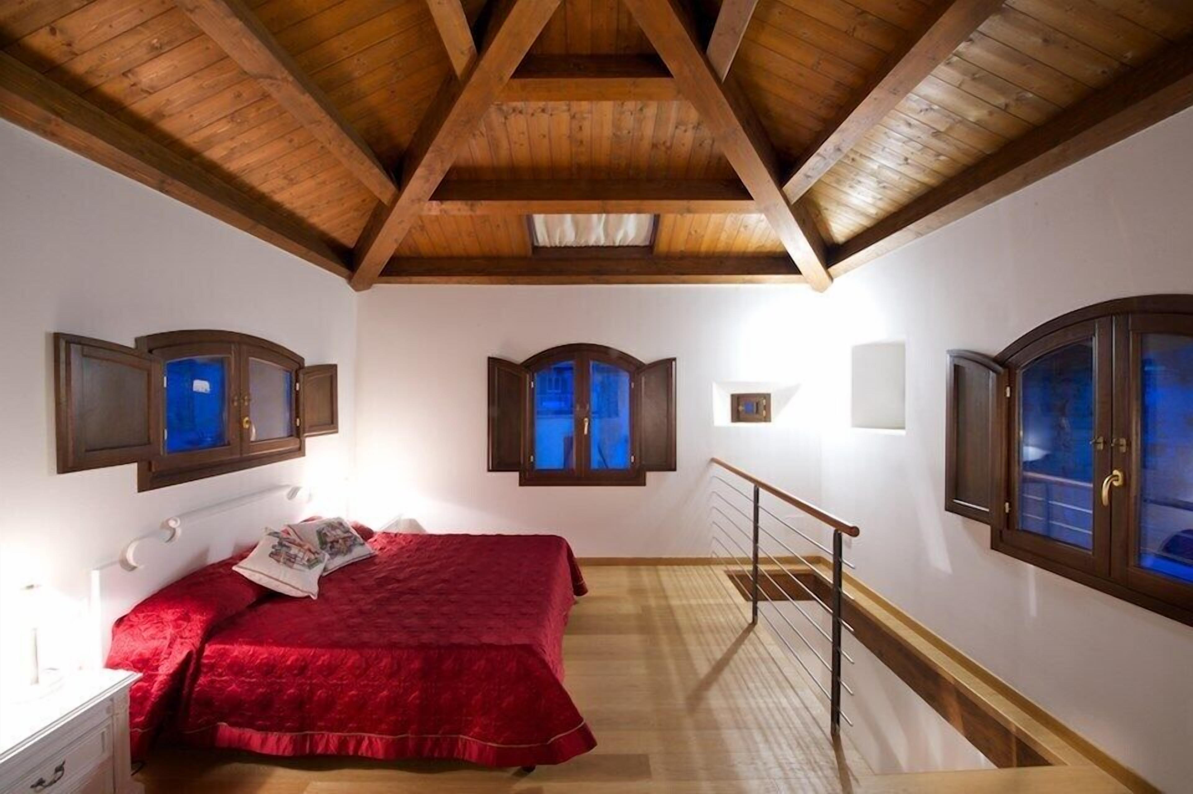 Een slaapkamer met een houten plafond, een bed met een rode sprei en twee ramen met houten kozijnen.