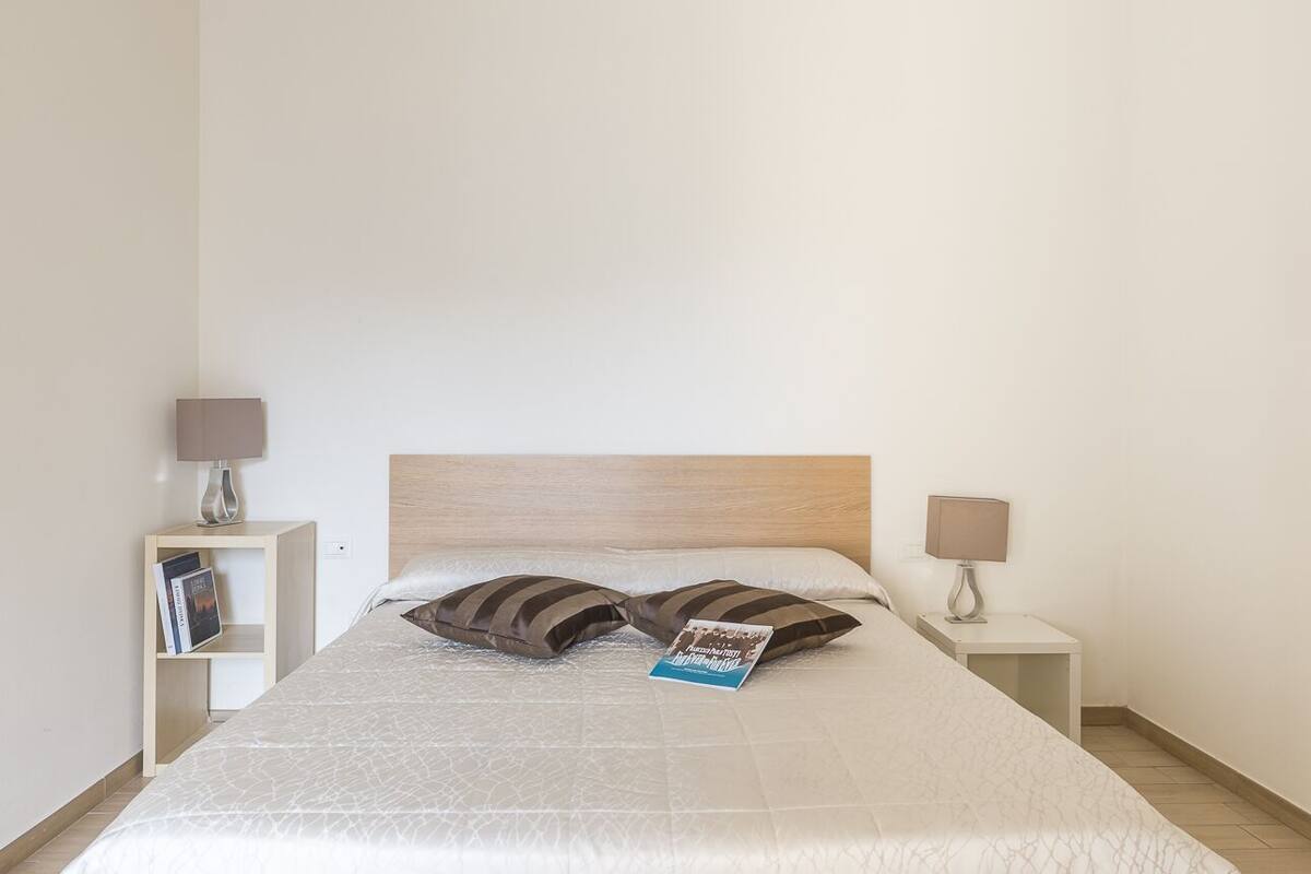 Un lit bien fait, avec une tête de lit en bois, deux oreillers et un magazine posé sur le lit. Il y a deux tables de chevet avec des lampes et une petite étagère avec des livres.