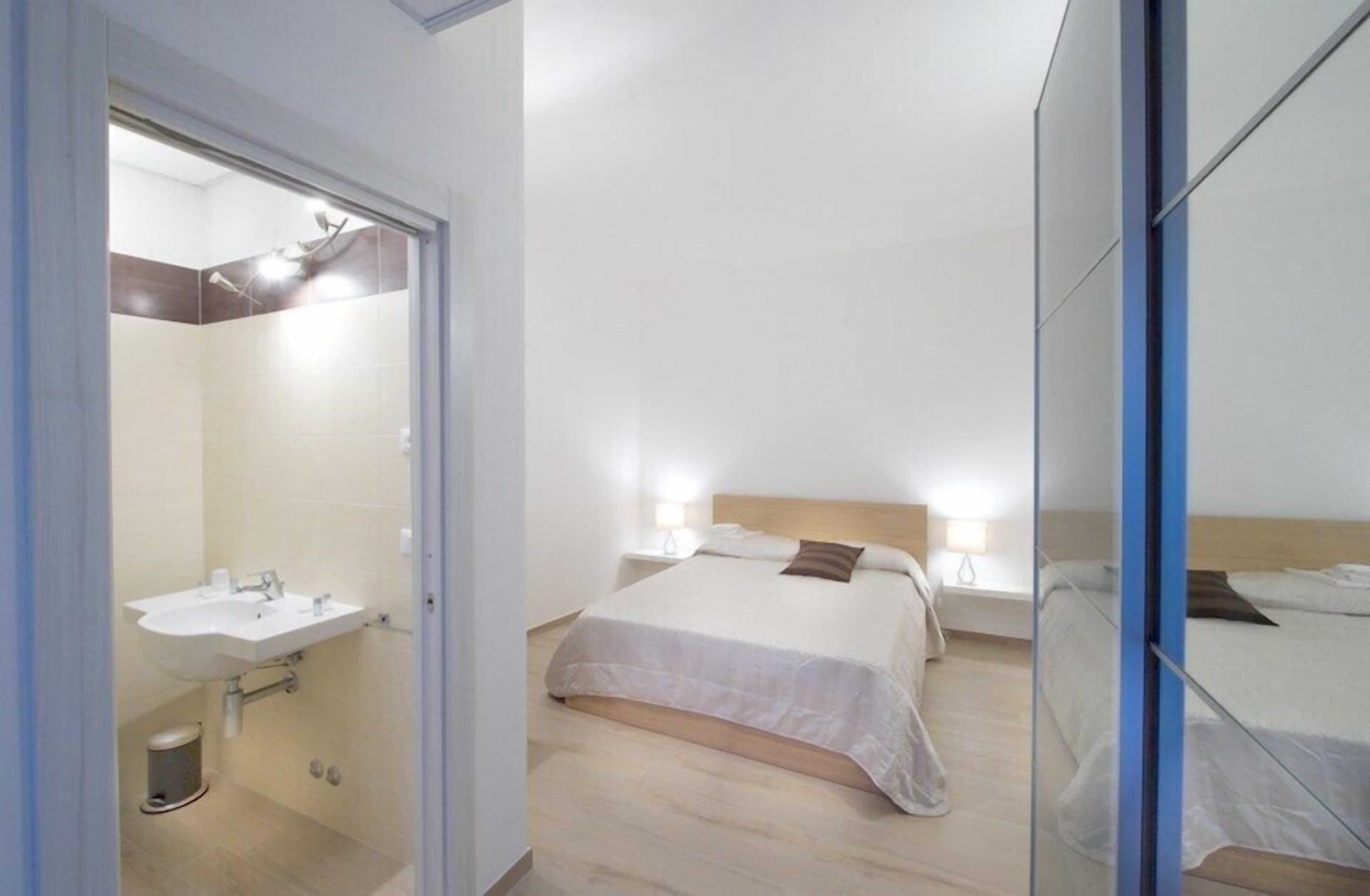 A modern hotel room with a large bed, bedside tables with lamps, a bathroom with a sink, and a mirror.