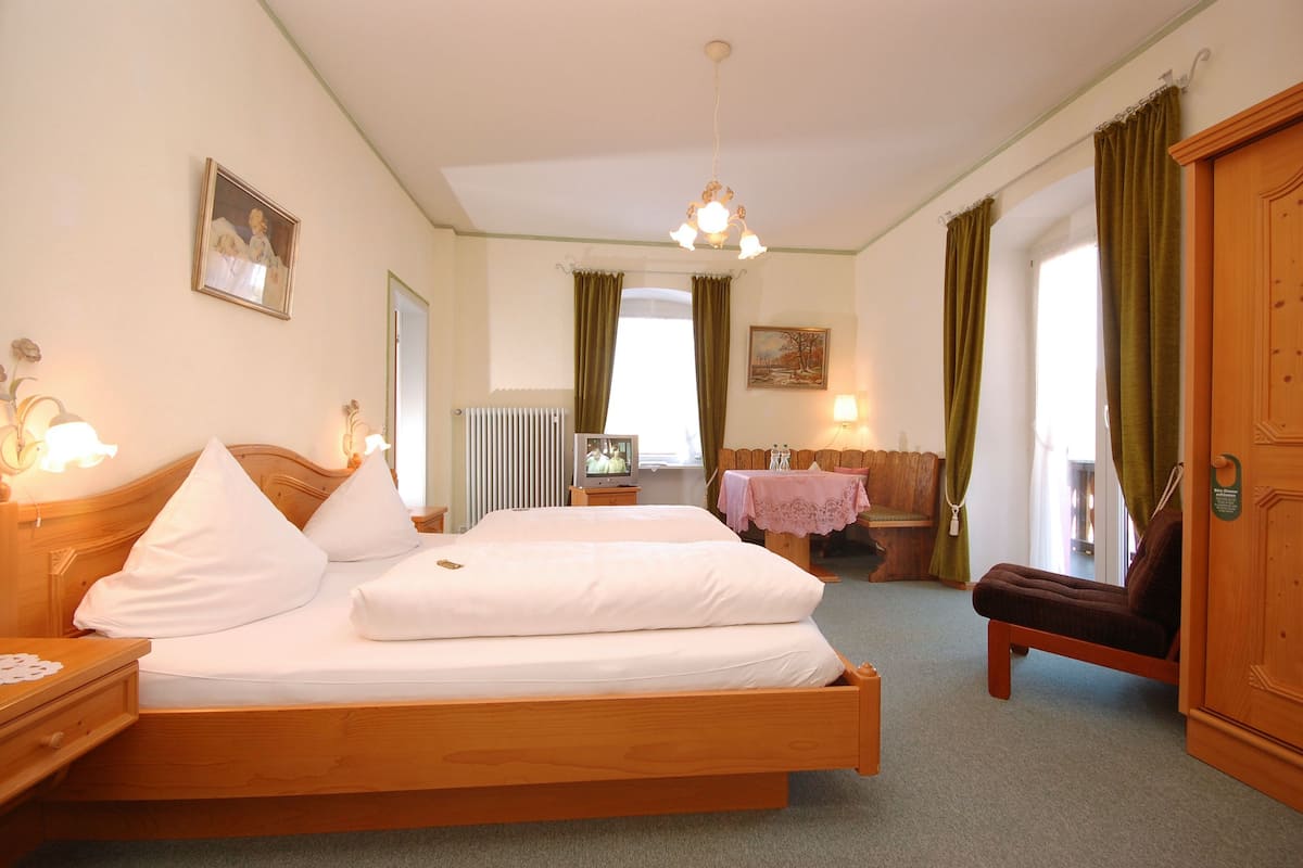 Et hotelværelse med en træseng, et natbord, en stol, et bord dækket af en dug, en radiator og et billede på væggen.
