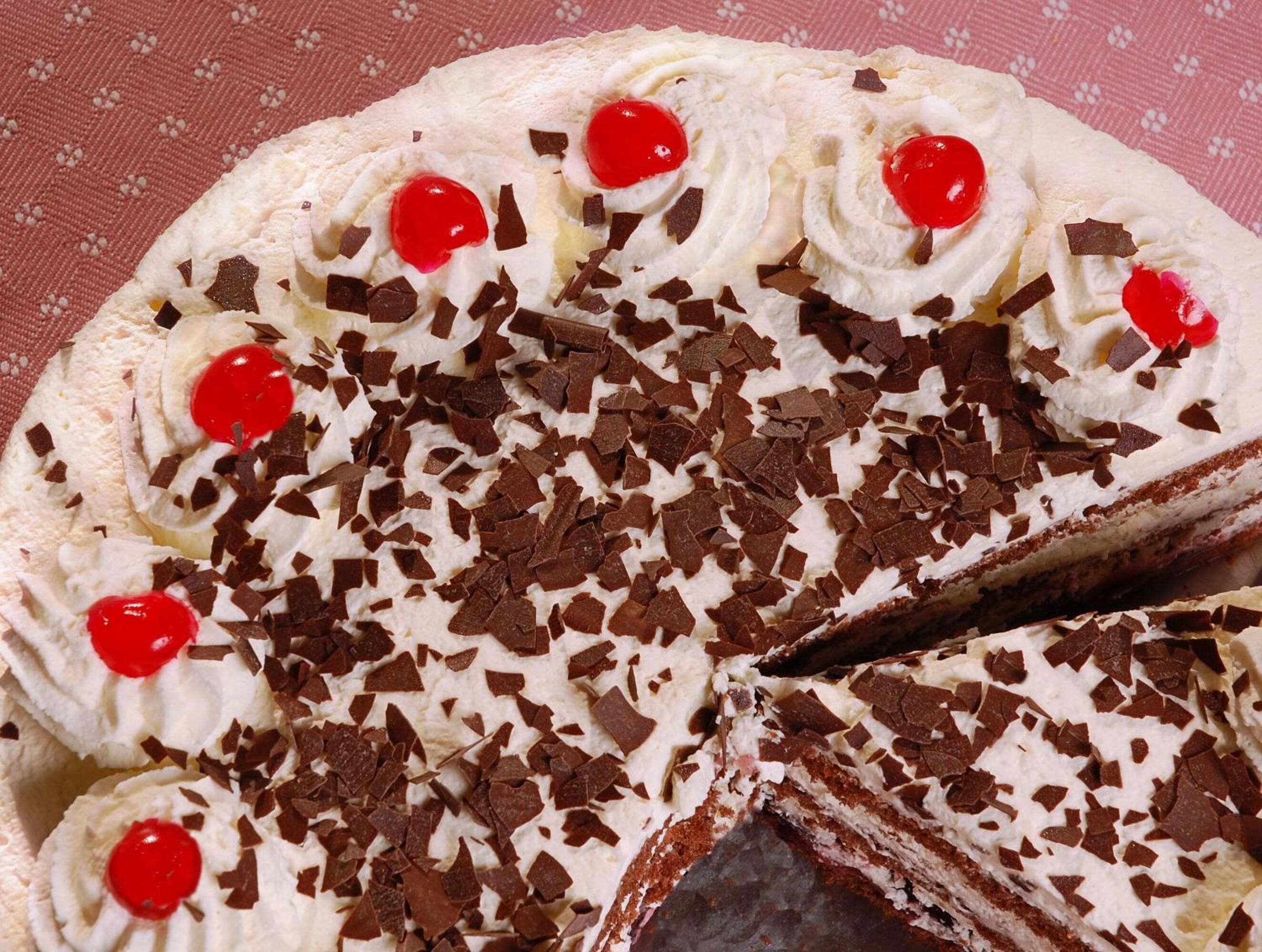 Un pastel redondo con crema batida, virutas de chocolate y cerezas por encima.