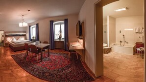 Ein modernes Hotelzimmer mit Holzboden, einer Essecke mit Tisch und Stühlen, einem Sofa, einem großen Teppich und einem Badezimmer mit Badewanne und Dusche.