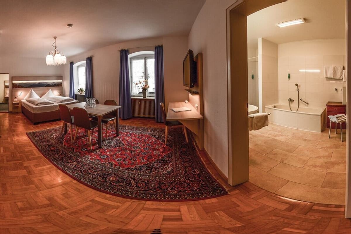 Et moderne hotelværelse med trægulv, et spiseområde med bord og stole, en sofa, et stort tæppe og et badeværelse med badekar og bruser.
