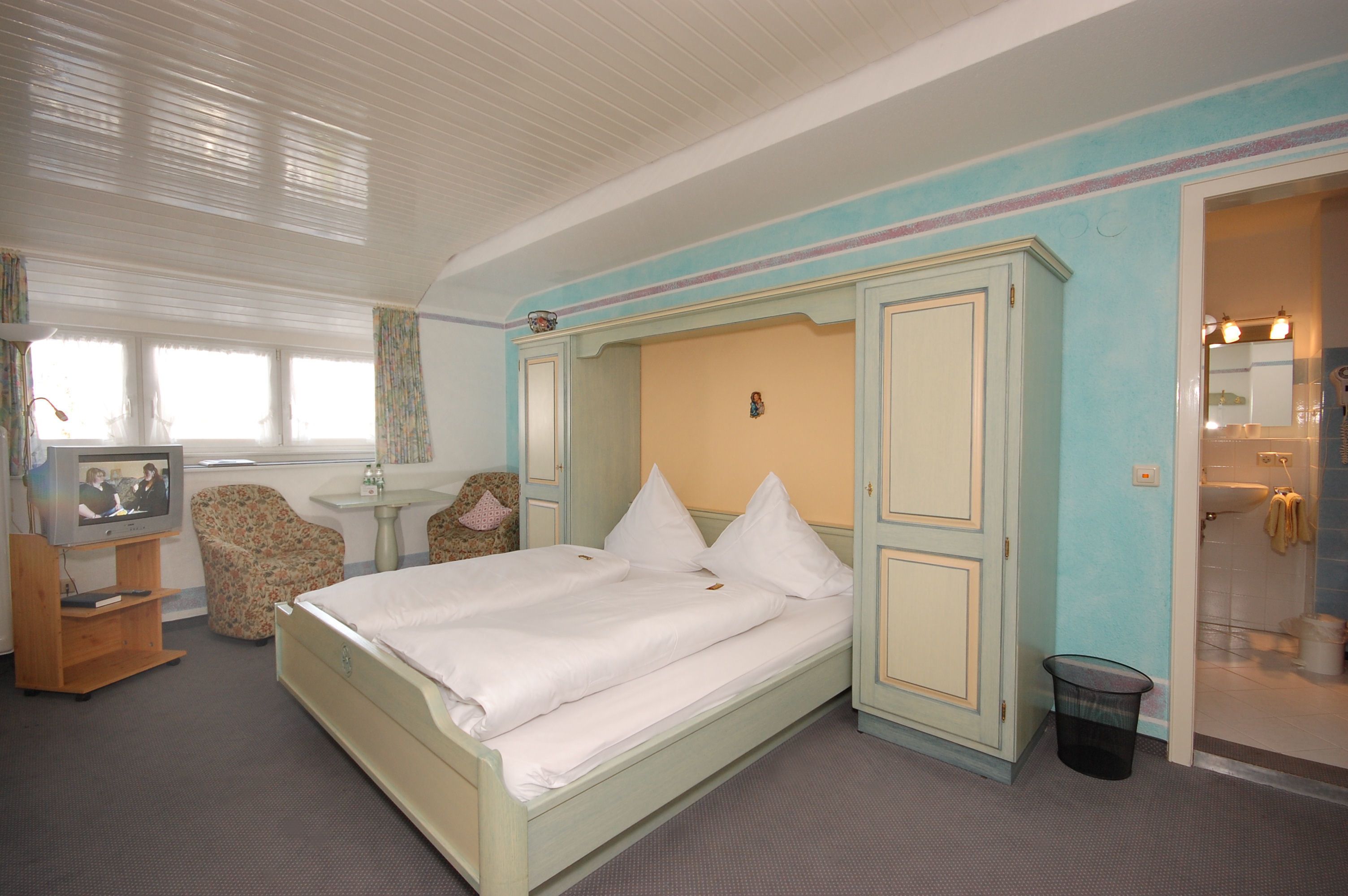 A hotel room with a bed, a television, a chair, a small table, and a bathroom visible through an open door.