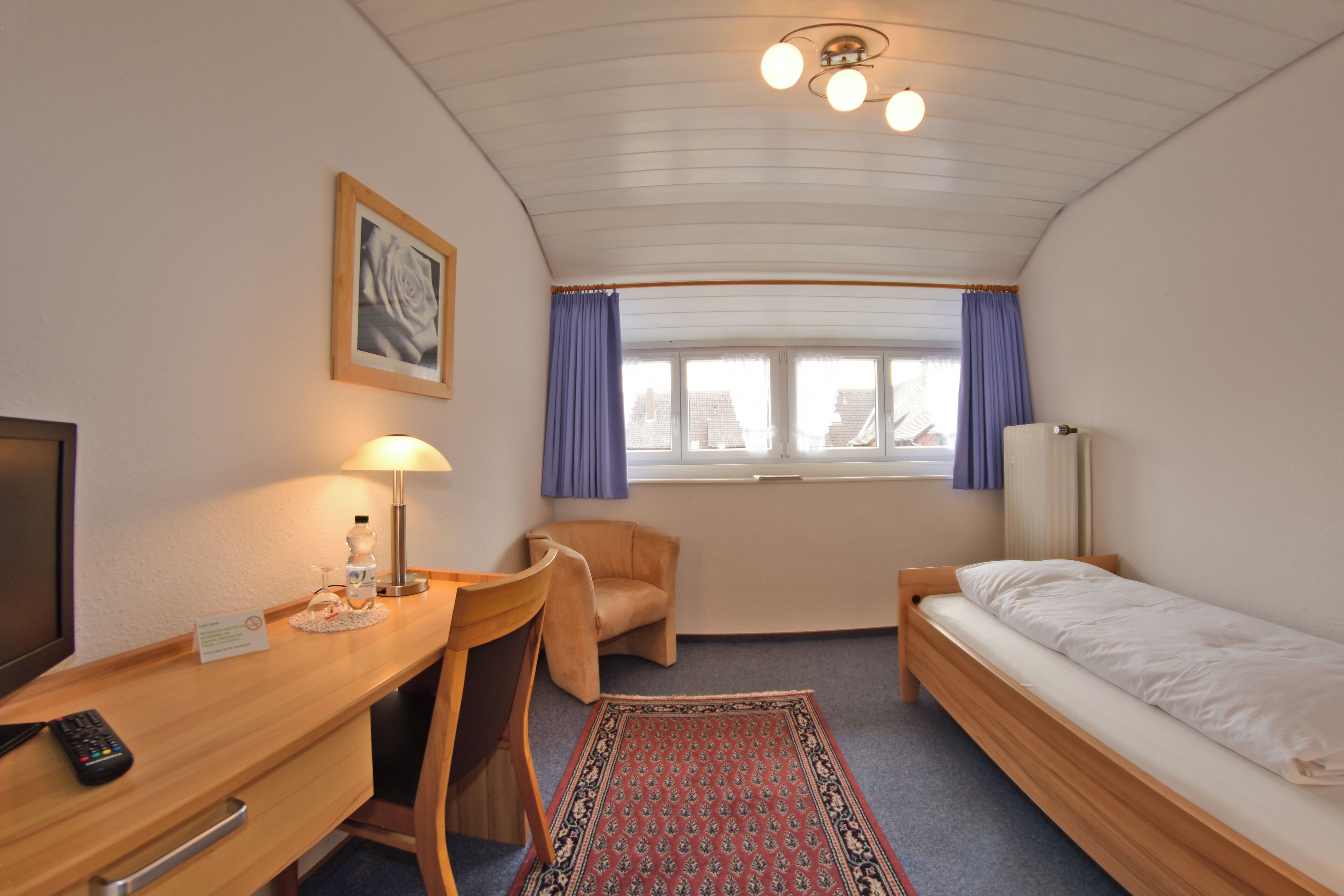 Habitación de hotel pequeña con una cama individual, un escritorio con lámpara, una silla, una mesita y un televisor.