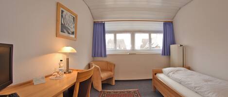 Habitación de hotel pequeña con una cama individual, un escritorio con lámpara, una silla, una mesita y un televisor.