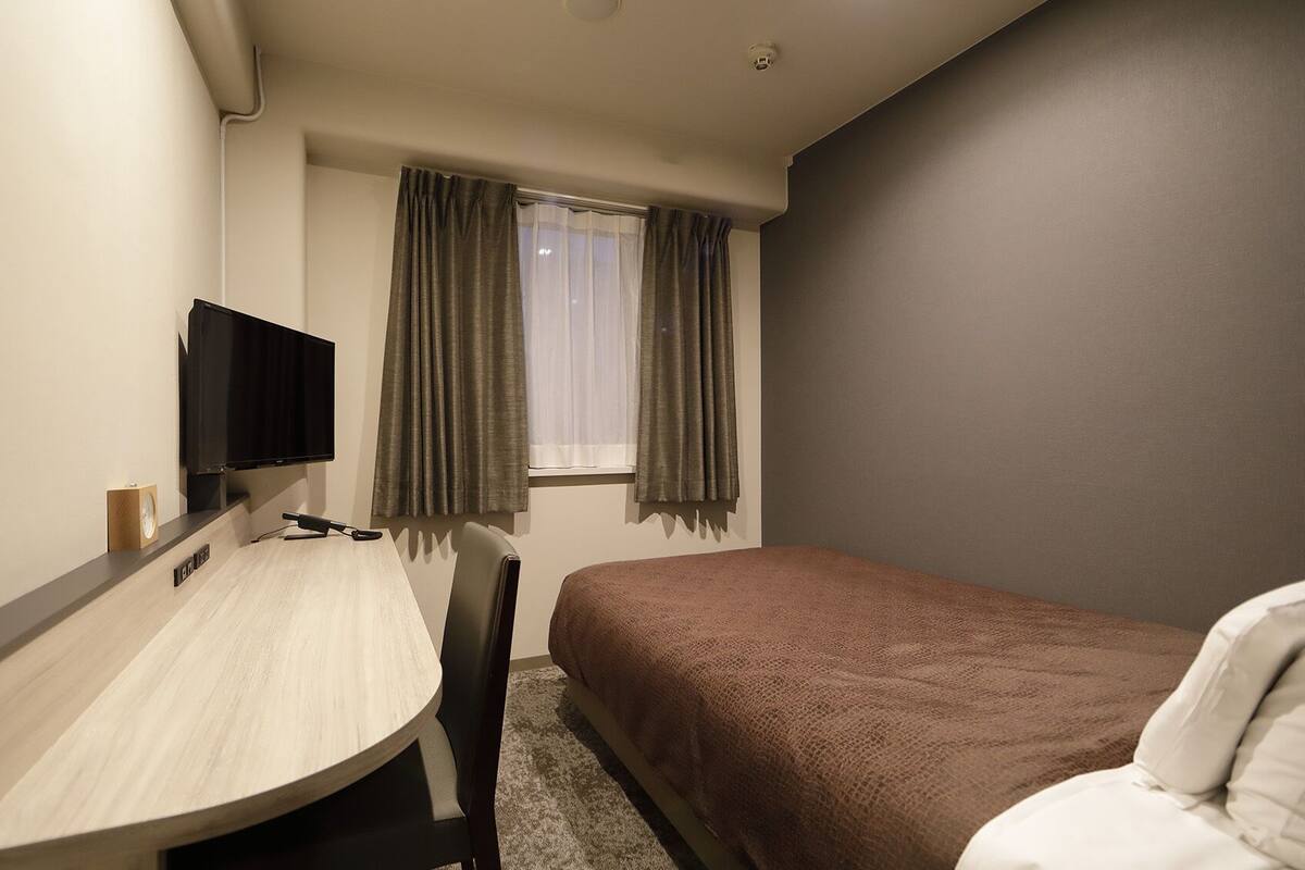 A hotel room with a bed, a desk with a chair, a television, and a window with curtains.