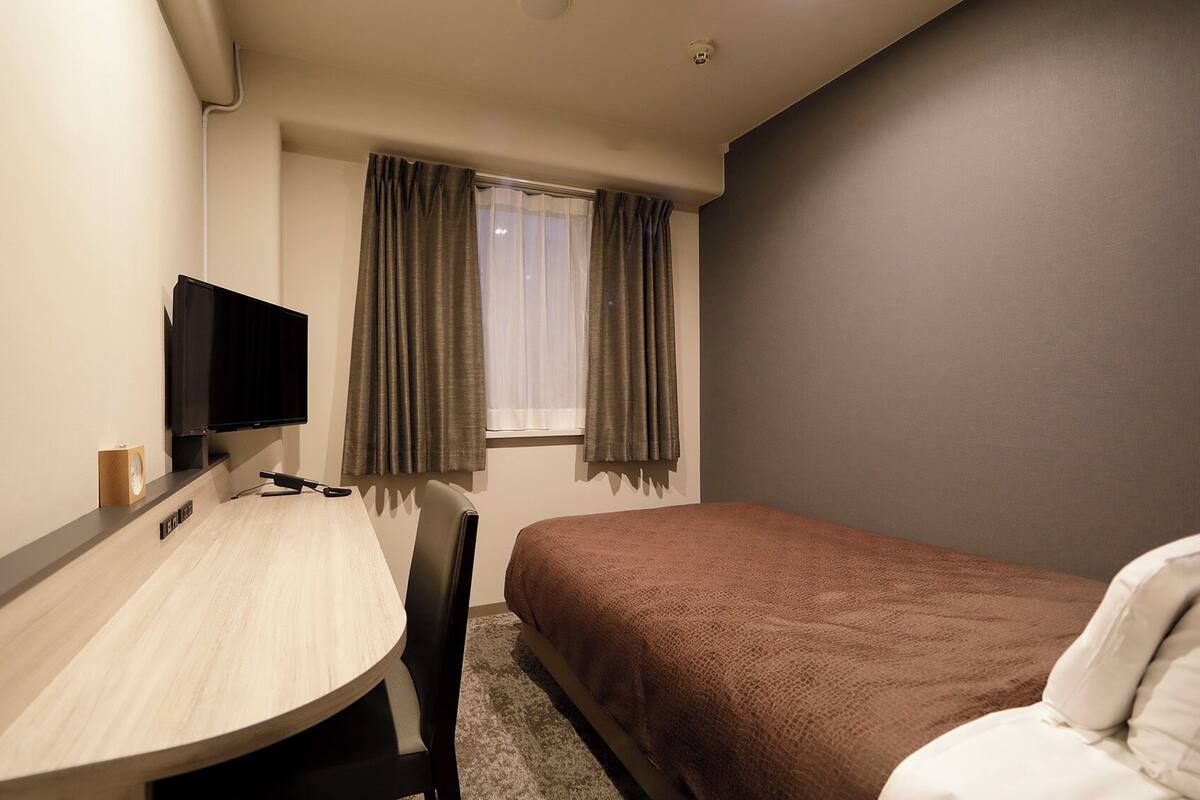 A hotel room with a bed, a desk with a chair, a television, and a window with curtains.