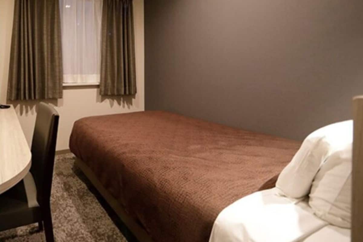A single bed with a brown cover, a chair, a window with curtains, and a plain wall.