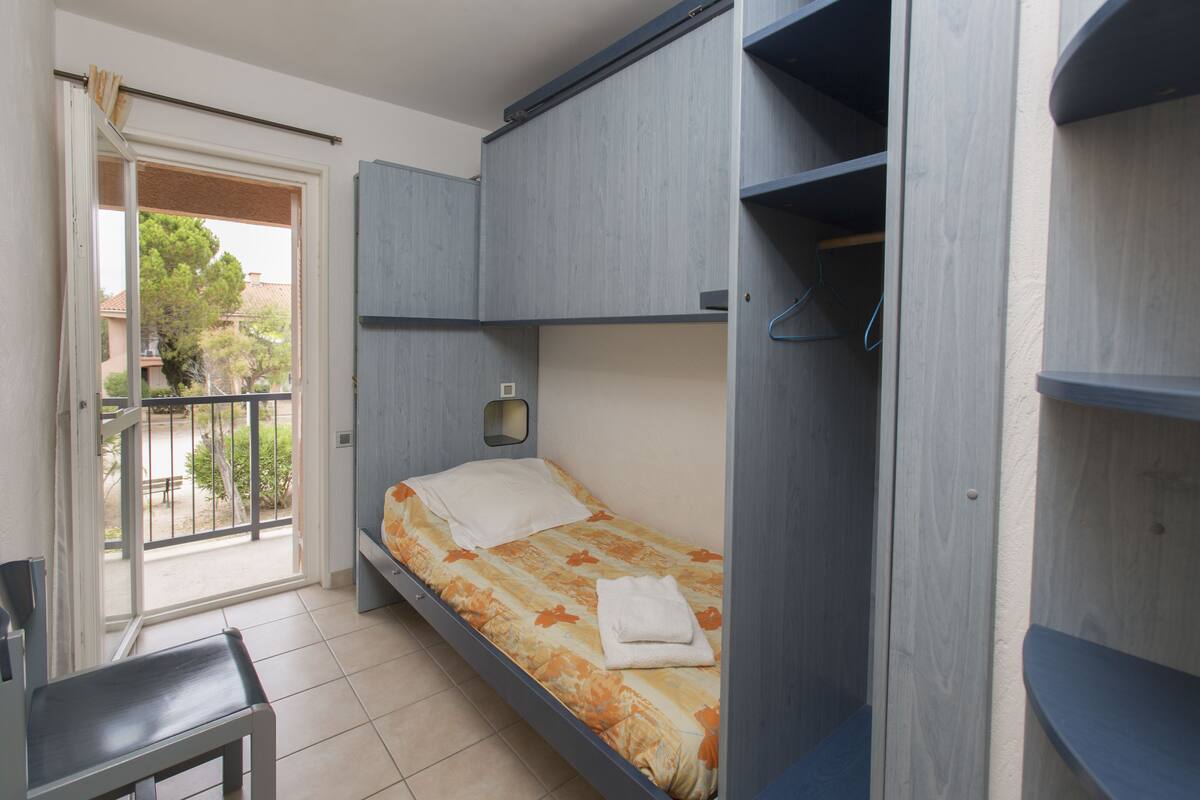 Une petite chambre avec un lit simple, une fenêtre, une chaise et une porte donnant sur un balcon.