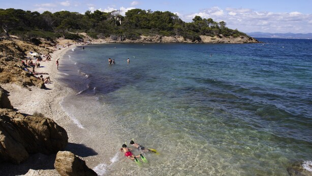 A beach with clear water, rocky shore, and a forested hill in the background. - Vacances Passion La Grande Bastide (Le Lavandou)