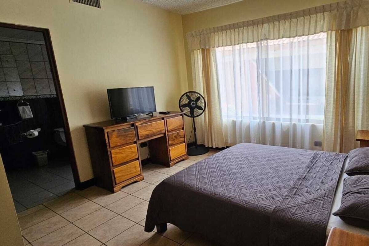 A bedroom with a bed, a TV, a fan, and a window with curtains.
