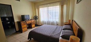 A bedroom with a bed, a TV, a fan, and a window with curtains. - Hotel Resort Obelisco (La Asunción)