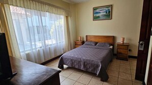 A bedroom with a bed, a TV, a wooden nightstand, a bedside table, and a painting on the wall. - Hotel Resort Obelisco (La Asunción)