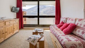 A room with a wooden cabinet, a sofa with pink pillows, a small table with newspapers, and a television on the wall.