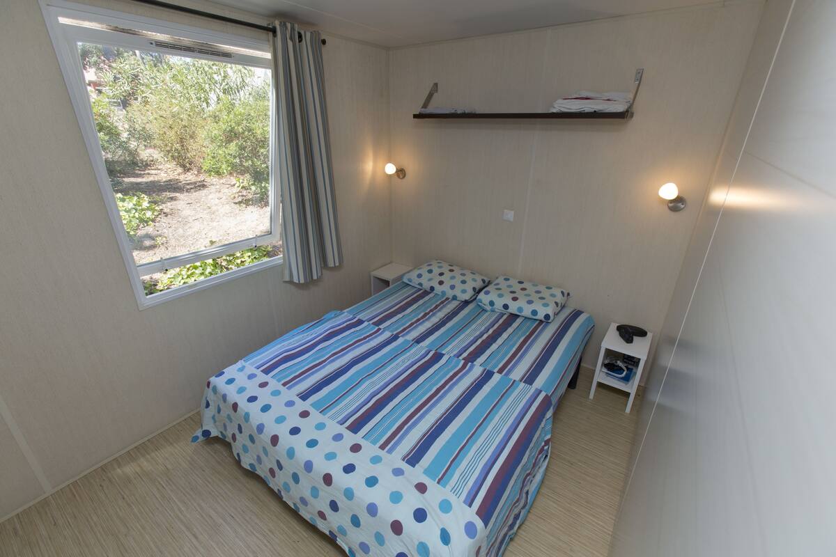 A small, neatly arranged bedroom with a single bed, a bedside table, a nightstand, a window with curtains, and a shelf above the bed.