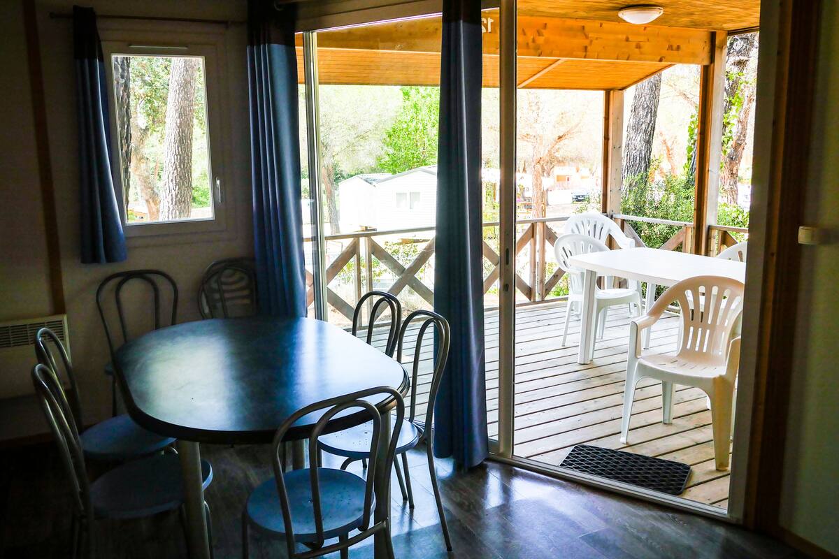 A room with a wooden table and chairs, a window with curtains, and a view of a deck with outdoor furniture.