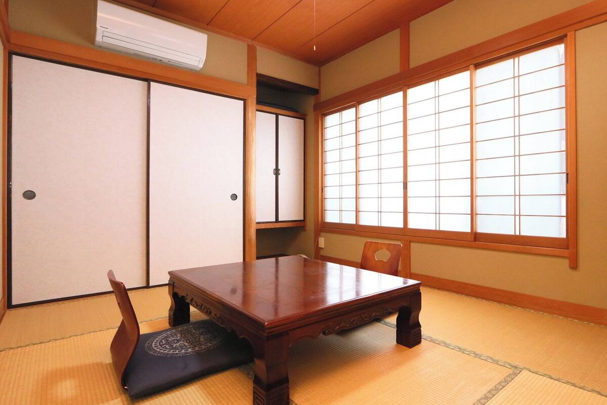A traditional Japanese room with tatami flooring, a low wooden table, a television on a stand, sliding doors, and a large window with shoji screens.