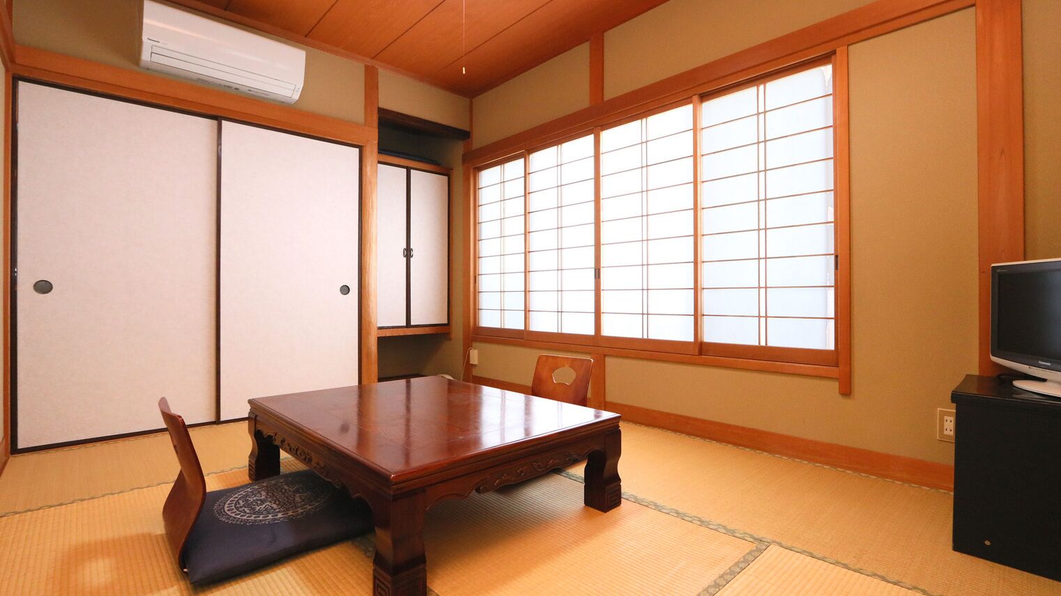 Een traditionele Japanse kamer met tatami vloerbedekking, een lage tafel, een stoel, schuifdeuren en een tv op een standaard.