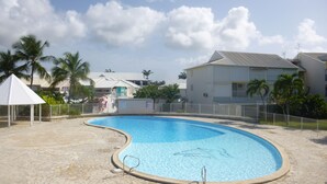 Una piscina de borde curvo, rodeada por una terraza de piedra. Se observan palmeras y un edificio blanco con techo gris al fondo.